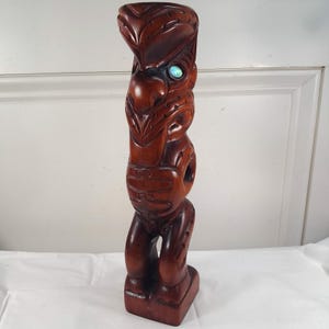 May include: A tall, dark brown wooden carving featuring intricate, stylized relief patterns across its surface. The figure has a prominent, curved head and a single, circular, iridescent abalone shell inlay set into the eye socket area.
