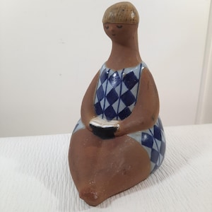 May include: A terracotta ceramic figurine of a seated person with a smooth, unglazed brown finish. The figure wears a blue and white diamond-patterned garment and holds a small, open book with black and white pages in their lap.