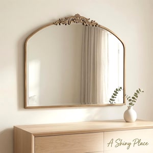 May include: An arched wall mirror with a gold-toned frame featuring an ornate, carved scrollwork crest at the top center. The mirror has a smooth, reflective glass surface and is mounted above a light-colored wooden dresser with the text A Shiny Place.