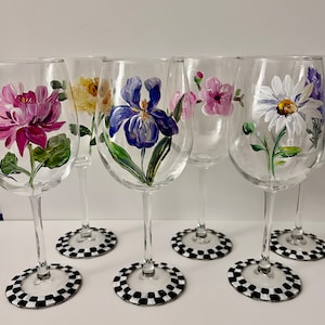 May include: Set of six clear glass wine glasses with hand-painted floral designs. Each glass features a different flower, including a pink dahlia, yellow rose, purple iris, pink flower, white daisy, and purple lavender. The glasses have black and white checkered bases.