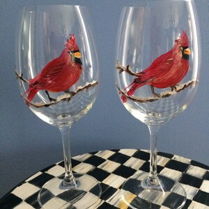 May include: Two clear wine glasses with painted red cardinals on a branch with snow. The glasses are on a black and white checkered surface.