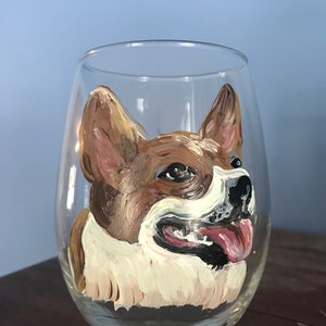 May include: A hand-painted wine glass featuring a portrait of a brown and white dog with a pink tongue sticking out. The dog's eyes are brown and the glass is clear.