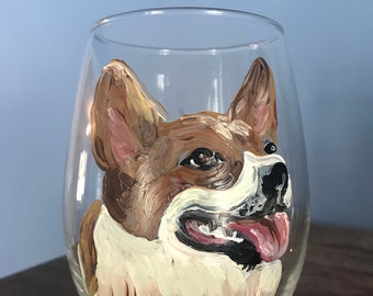 May include: A hand-painted wine glass featuring a portrait of a brown and white dog with a pink tongue sticking out. The dog's eyes are brown and the glass is clear.