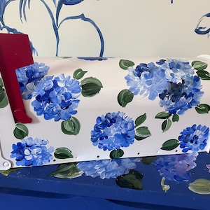 May include: White mailbox with a red flag, painted with blue hydrangea flowers and green leaves.