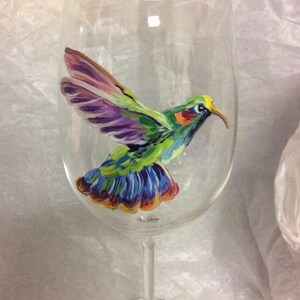 May include: A clear glass wine glass with a hand-painted hummingbird design. The hummingbird is green, purple, blue, and yellow.