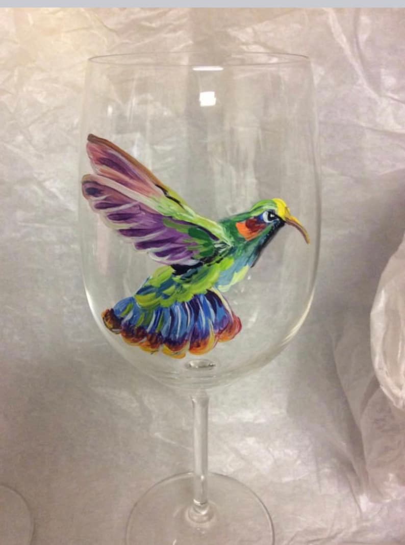 May include: A clear glass wine glass with a hand-painted hummingbird design. The hummingbird is green, purple, blue, and yellow.