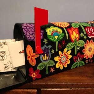 May include: A black metal post box featuring a hand-painted floral pattern in vibrant shades of red, orange, yellow, purple, and green. A bright red flag is attached to the side. Inside the post box are several white greeting cards with black ink.
