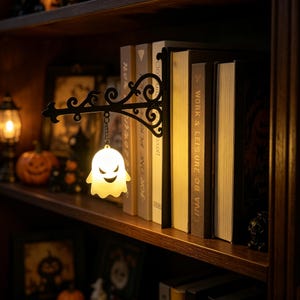 May include: A black metal bookend featuring an ornate, scrollwork bracket that extends outward. A small, glowing white ghost-shaped light hangs from the bracket by a short chain, illuminating the spines of several books on a wooden shelf.