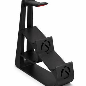 May include: A black gaming accessory stand featuring a tall vertical support with a red accent on the top hook and two lower tiers. The structure includes cut-out Xbox logos on the front panels, designed to hold controllers and a headset.