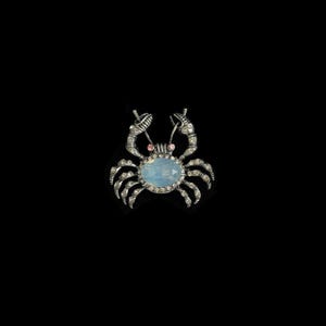 Op de afbeelding: Een zilverkleurige krab-broche met een centrale ovaalvormige iriserende lichtblauwe steen. Het lichaam en de poten van de krab zijn ingelegd met kleine doorzichtige strass-steentjes en het heeft twee kleine roze steentjes als ogen op een zwarte achtergrond.