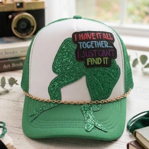 May include: A white and green trucker hat featuring a large green glittery bow appliqué and a black patch with the text I HAVE IT ALL TOGETHER... I JUST CAN'T FIND IT. The cap includes a gold-toned chain detail across the base of the front panel.