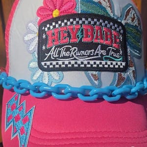 May include: A pink and white trucker hat featuring a black rectangular patch with the text HEY BABE All The Rumors Are True. The hat is decorated with embroidered flower patches, a blue plastic chain link trim, and a blue and pink lightning bolt.