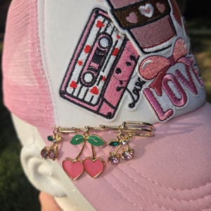 May include: A light pink trucker hat featuring a white front panel with embroidered patches of a coffee cup, a cassette tape, and the word LOVE. A gold-toned safety pin is attached to the brim, adorned with three dangling cherry charms.
