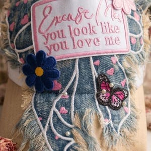 May include: A distressed denim baseball cap featuring a rectangular patch with the text Excuse Me you look like you love me in pink embroidery. The cap is decorated with a pink flower, a blue flower, a butterfly patch, and small white beads.