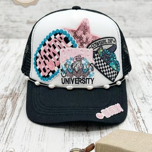 May include: A black and white trucker hat featuring a pink sequined star, a blue and black checkered patch with pink text, and a central patch reading COWGIRL UNIVERSITY with a graphic of a person in a hat. A string of white pearls accents the brim.