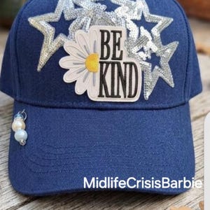May include: A navy blue baseball cap featuring silver sequined star appliques and a central patch with a white daisy and the text BE KIND. A small charm with three round beads in white and blue tones is attached to the left side of the brim.