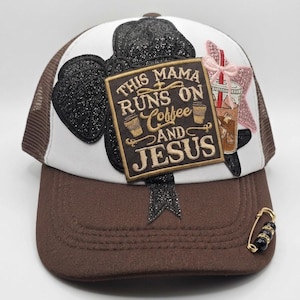 May include: A brown and white trucker hat featuring a black glittery bow, a pink glittery star, and a rectangular patch with the text "THIS MAMA RUNS ON Coffee AND JESUS". A gold safety pin with black and gold beads is attached to the brown brim.