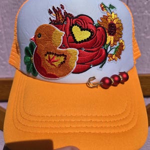 May include: An orange and white trucker hat featuring an embroidered patch of a yellow duck, a large red rose with a yellow heart, and a sunflower. The design includes small rhinestones and a safety pin accessory with three red beads on the brim.