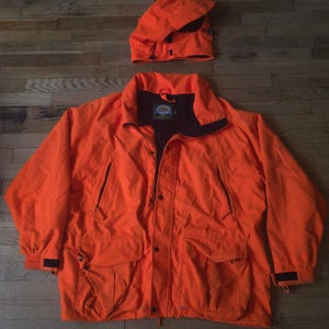 May include: A bright orange Cabela's jacket with a detachable hood, featuring a black interior lining, a front zipper closure with snap buttons, two large lower cargo pockets, and two vertical zippered chest pockets with adjustable wrist cuffs.
