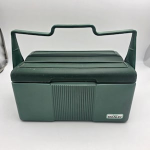 May include: A dark green plastic lunch box with a rectangular shape and a large, integrated top handle. The front features a vertical ribbed texture in the center and a small white rectangular label in the bottom right corner with the text Stanley.