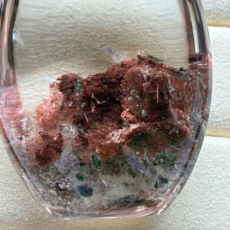 May include: A clear, oval-shaped quartz stone containing internal mineral inclusions. The interior features a dense cluster of deep reddish-brown, earthy deposits mixed with small, dark green mineral flecks embedded within the transparent crystal structure.