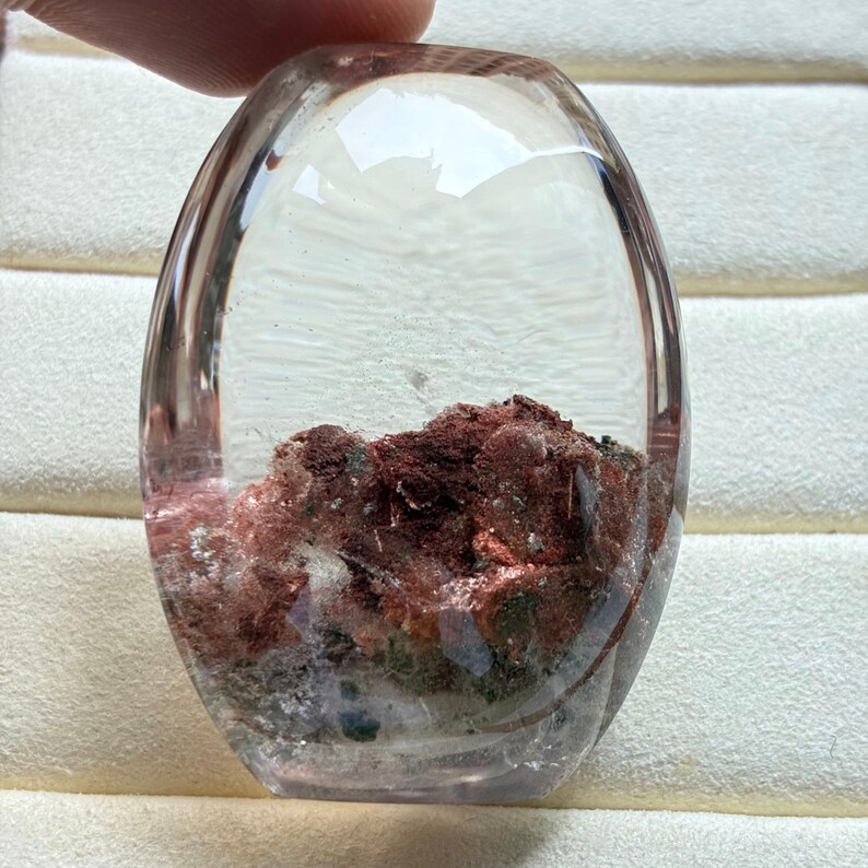 May include: A polished, oval-shaped clear quartz gemstone featuring internal inclusions of deep red and dark green mineral deposits. The stone has a smooth, rounded surface that magnifies the textured, earthy cluster of minerals trapped inside the crystal.