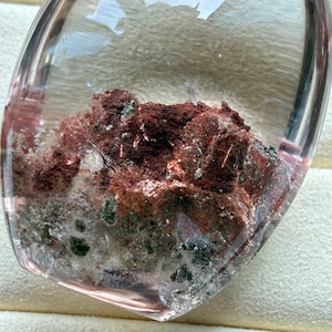 May include: A polished, teardrop-shaped clear quartz gemstone containing internal mineral inclusions. The stone features prominent patches of deep red, rust-coloured deposits alongside smaller, dark green mineral flecks embedded within the transparent, glassy structure of the crystal.