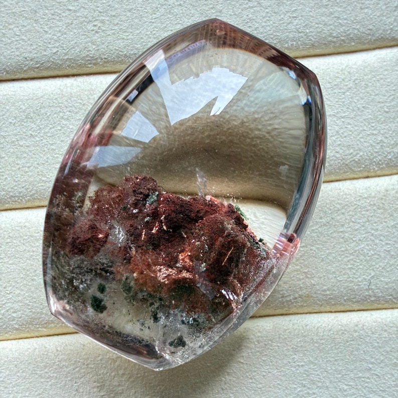 May include: A polished, shield-shaped clear quartz gemstone featuring prominent internal inclusions of deep reddish-brown mineral deposits and small dark green flecks, encased within a smooth, transparent, and faceted crystalline structure.