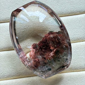 May include: A polished, shield-shaped clear quartz gemstone featuring prominent internal inclusions of deep red and rust-coloured mineral deposits concentrated in the lower section, with a smooth, transparent, and faceted surface finish.