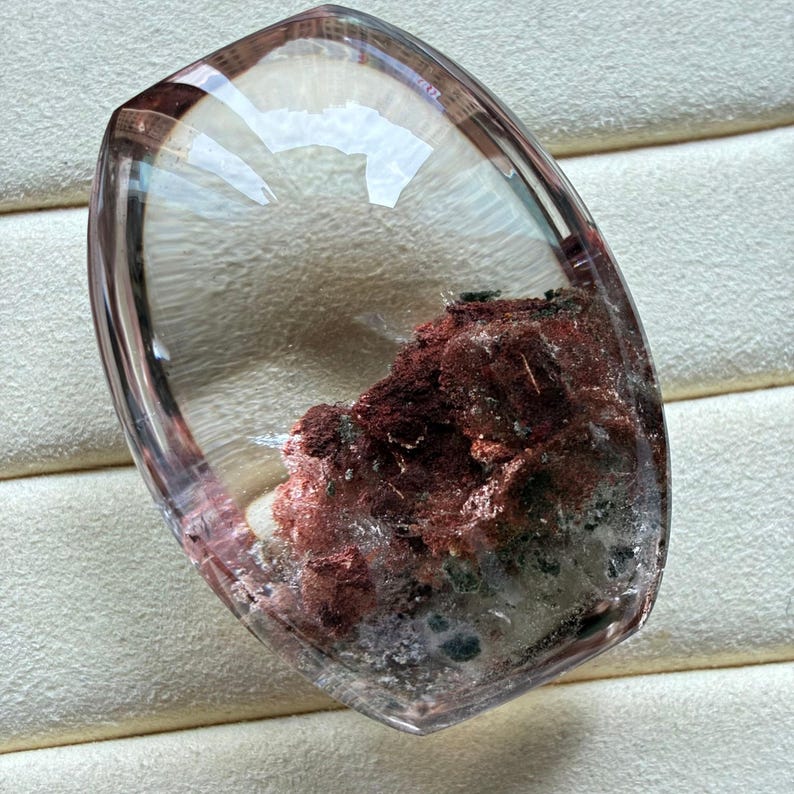 May include: A polished, shield-shaped clear quartz gemstone featuring prominent internal inclusions of deep red and rust-coloured mineral deposits concentrated in the lower section, with a smooth, transparent, and faceted surface finish.
