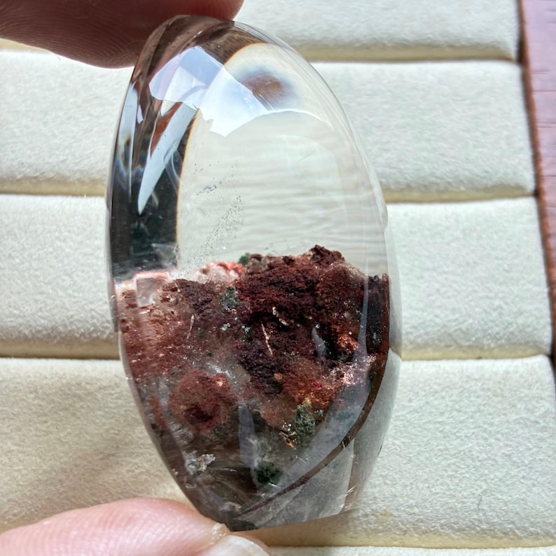 May include: A polished, oval-shaped clear quartz crystal cabochon featuring internal inclusions of deep red and dark brown mineral deposits. The transparent stone has a smooth, rounded surface that reveals the textured, earthy red and brown mineral formations trapped within the clear crystalline structure.