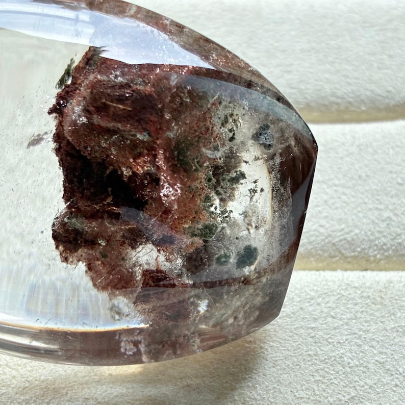 May include: A polished, clear quartz crystal point featuring prominent internal inclusions of deep reddish-brown and dark green mineral deposits. The transparent stone has a smooth, rounded surface that reveals the complex, textured mineral formations trapped within the crystalline structure.