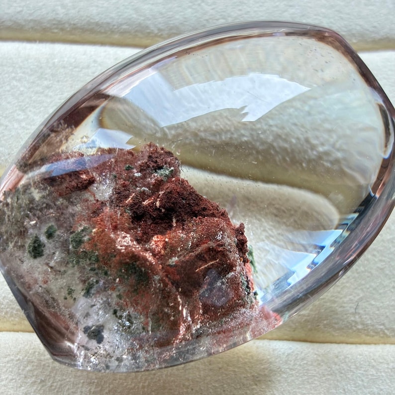 May include: A polished, oval-shaped clear quartz crystal featuring internal inclusions of deep red, rust-coloured, and dark green mineral deposits. The stone has a smooth, rounded surface with a high-gloss finish that reflects light across its transparent body.