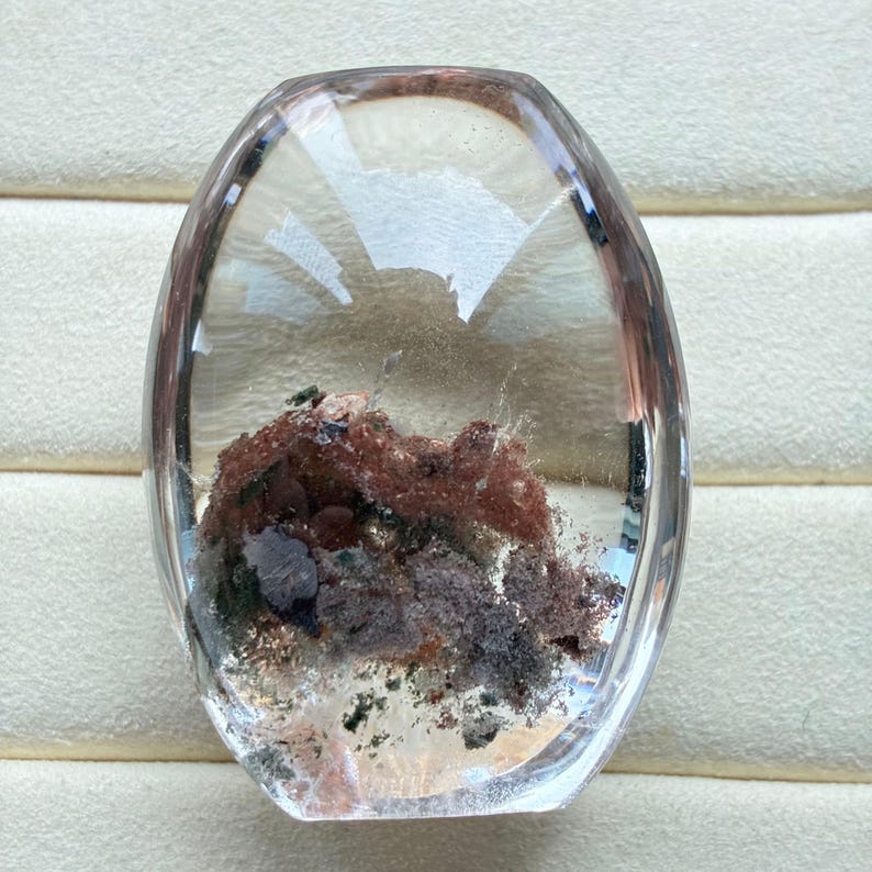 May include: A polished, oval-shaped clear quartz gemstone featuring internal mineral inclusions. The inclusions display a mix of earthy red, brown, and dark grey tones, creating a textured, landscape-like appearance within the transparent stone.