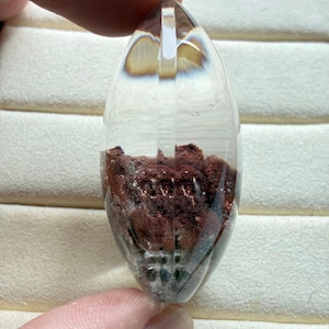 May include: A marquise-shaped clear quartz pendant featuring a prominent inclusion of deep reddish-brown mineral deposits at the base. The transparent stone has a smooth, polished surface and a faceted top, showcasing the internal texture of the earth-toned sediment trapped within the crystalline structure.