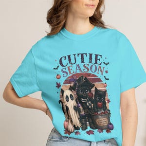 May include: A bright turquoise t-shirt featuring a graphic print with the text CUTIE SEASON. The design includes a ghost, a black cat dressed as a pirate, and a black cat in a bucket, surrounded by pumpkins, autumn leaves, and bats.