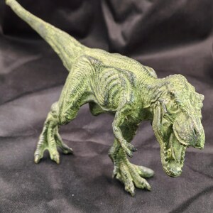 Puede incluir: Una figura de dinosaurio Tyrannosaurus Rex de color verde con un acabado texturizado y desgastado. La figura se muestra en una postura de pie, inclinada hacia adelante con la boca abierta, mostrando escamas detalladas y garras afiladas.