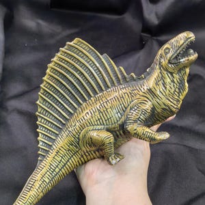 Op de afbeelding: Een Dimetrodon dinosaurusfiguur met een opvallend rugzeil met verticale ribbels. Het wezen is gekleurd in tinten olijfgroen en geel met een gestructureerd, geschubd oppervlak, een open bek en een lange, taps toelopende staart die naar buiten steekt.