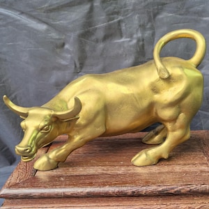 Puede incluir: Una figura de toro de color dorado en posición de carga con la cabeza baja y la cola curvada hacia arriba. El toro presenta un acabado metálico liso y está colocado sobre una base de madera rectangular con detalles tallados en los bordes.