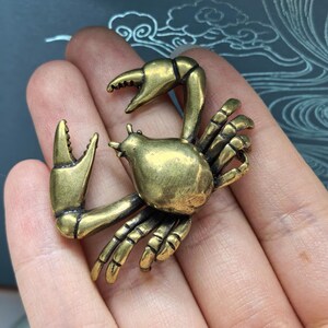 May include: A brass-coloured metal crab figurine with a smooth, polished finish. The crab is depicted with its two large claws raised upwards and several smaller legs tucked underneath its rounded body, showcasing a detailed, solid construction.