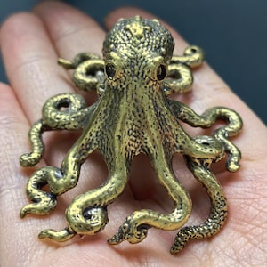 May include: A metallic gold-toned octopus figurine featuring a textured surface with intricate detailing on the head and multiple curved tentacles extending outward. The creature has two dark, prominent eyes and a rounded, bumpy mantle.