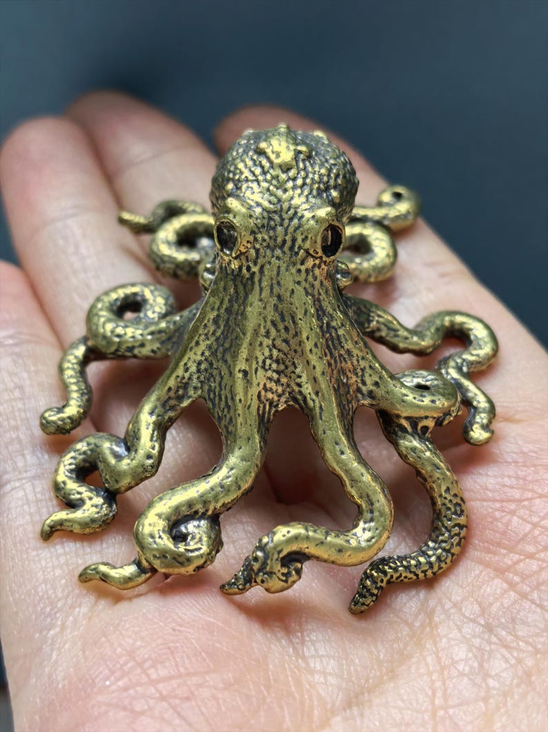 May include: A metallic gold-toned octopus figurine featuring a textured surface with intricate detailing on the head and multiple curved tentacles extending outward. The creature has two dark, prominent eyes and a rounded, bumpy mantle.