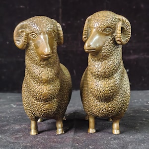 Puede incluir: Dos figuras de carnero de pie fabricadas en un material metálico de tono bronce. Cada carnero presenta patrones de lana texturizados y detallados en el cuerpo, cuernos curvados a los lados de la cabeza y rasgos faciales suaves y definidos.