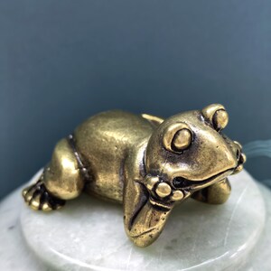 May include: A small, metallic, brass-colored frog figurine in a resting position. The frog has a smooth, polished surface with visible dark patina in the crevices, large eyes, and front limbs tucked underneath its head in a relaxed pose.