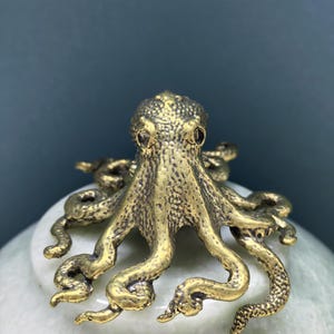 May include: A small, metallic, gold-toned octopus figurine with a textured, bumpy surface. The octopus has a rounded head and eight curved, sprawling tentacles that extend outward from the center, showcasing intricate detailing on the skin.