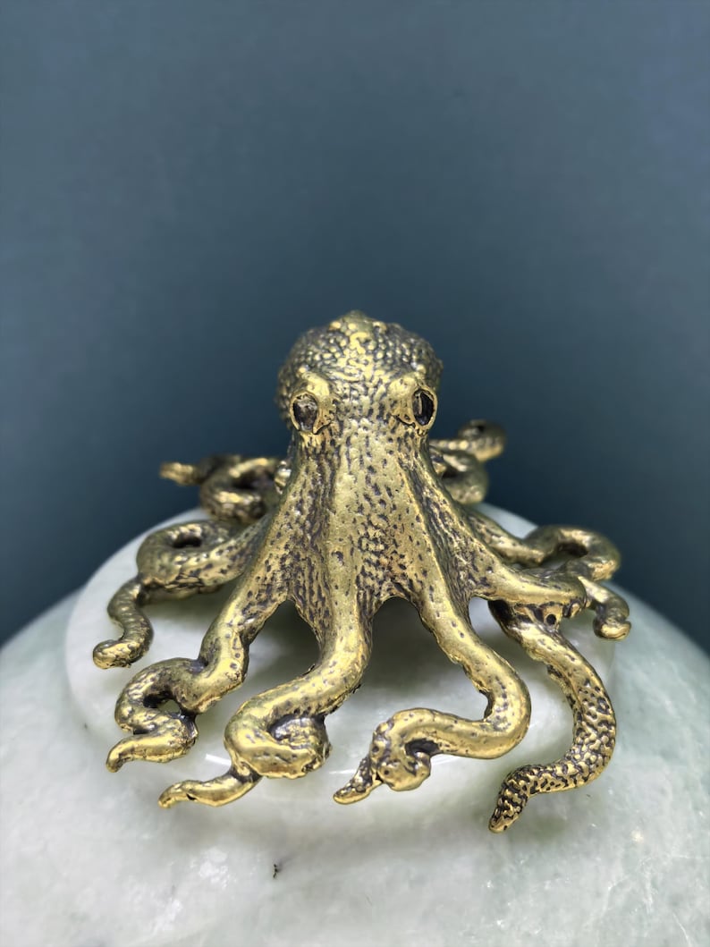 May include: A small, metallic, gold-toned octopus figurine with a textured, bumpy surface. The octopus has a rounded head and eight curved, sprawling tentacles that extend outward from the center, showcasing intricate detailing on the skin.