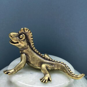 May include: A small, metallic, brass-colored lizard figurine with a textured, ridged back and a long, curved tail. The lizard is depicted in a standing position with its head turned slightly to the left, featuring detailed eyes and a small mouth.