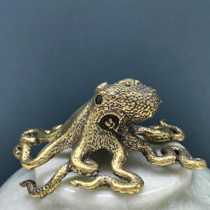 May include: A small, metallic, gold-toned octopus figurine with a textured, bumpy surface. The octopus has a rounded head and eight curved, sprawling tentacles that extend outward, showcasing intricate detailing on the skin and suction cups.