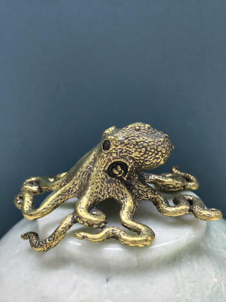 May include: A small, metallic, gold-toned octopus figurine with a textured, bumpy surface. The octopus has a rounded head and eight curved, sprawling tentacles that extend outward, showcasing intricate detailing on the skin and suction cups.