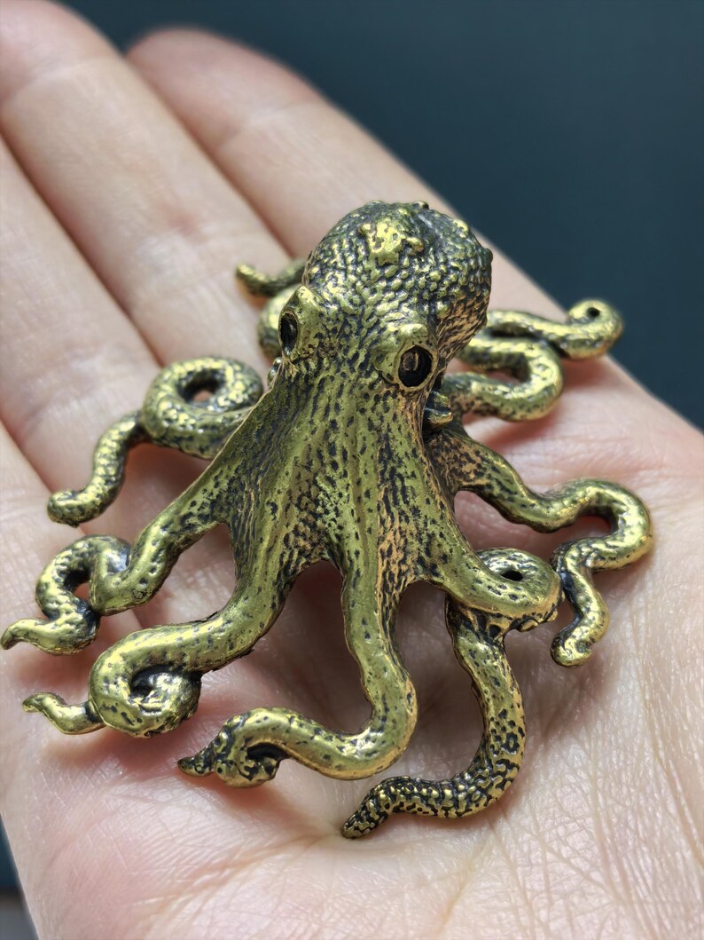 May include: A small, three-dimensional brass octopus figurine with a textured, mottled surface. The creature features a rounded head, two dark, recessed eyes, and eight curved, sprawling tentacles that extend outward in various directions.